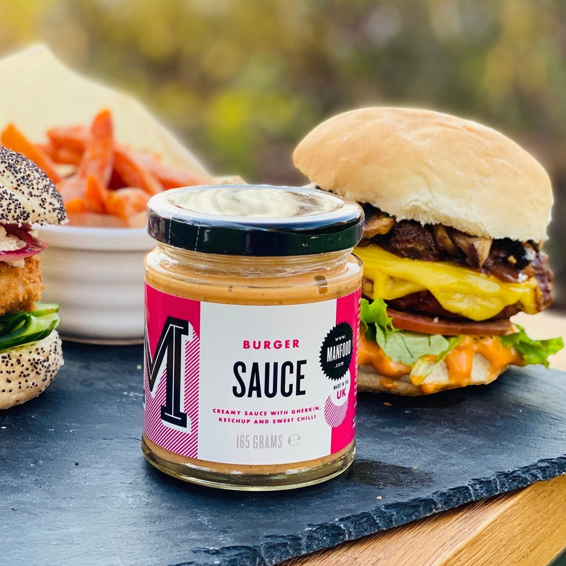 Manfood vegan burger sauce jar on a slate board next to a gourmet burger and sweet potato fries.